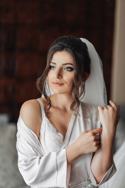 Brunette bride in a robe, posing for the camera, looking to the side. Open shoulders. Elegant hairstyle. Nice makeup. Voluminous veil. Morning of the bride. Beautiful hands of the bride - Photo, Image