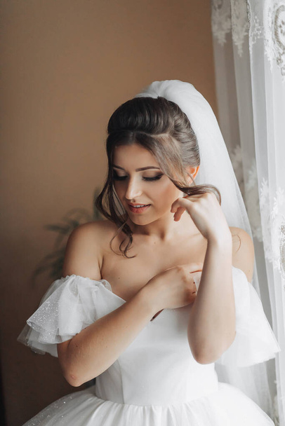 A brunette bride in a white dress holds her gorgeous curls and looks down. Open shoulders. Beautiful hands. Long veil. Morning of the bride. - Foto, Imagem