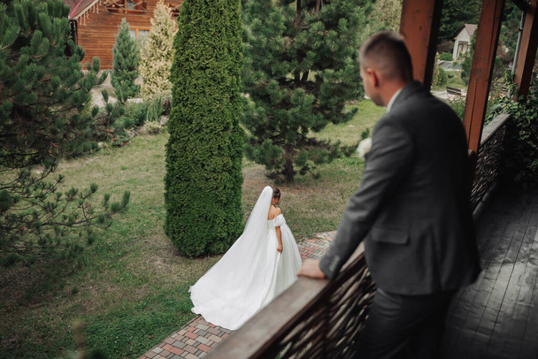 Wedding photo. The bride in a voluminous white dress and a long veil walks in the garden along a stone path, the groom stands on the balcony and looks at her. View from above. Beautiful makeup - Foto, immagini