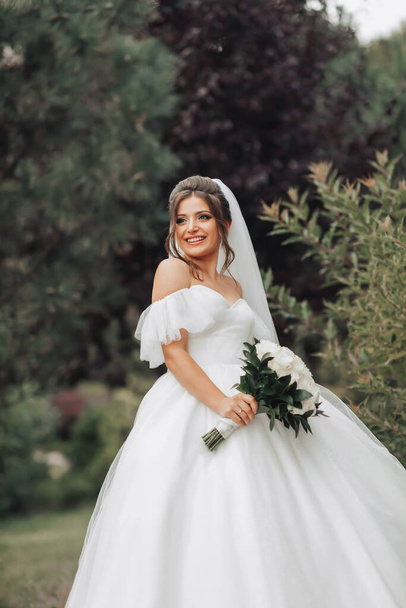 Wedding photo. The bride in a voluminous white dress and a long veil, smiling, twirling with a bouquet of white roses, holding her dress. Portrait of the bride. Beautiful makeup and hair. - Foto, afbeelding