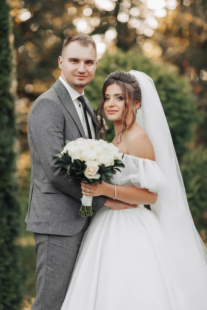 Portrait of the bride and groom in nature. Cropped photo. The bride and groom pose, hugging and smiling, against the background of conifers. The bride in a long dress with a bouquet of roses. - Fotografie, Obrázek