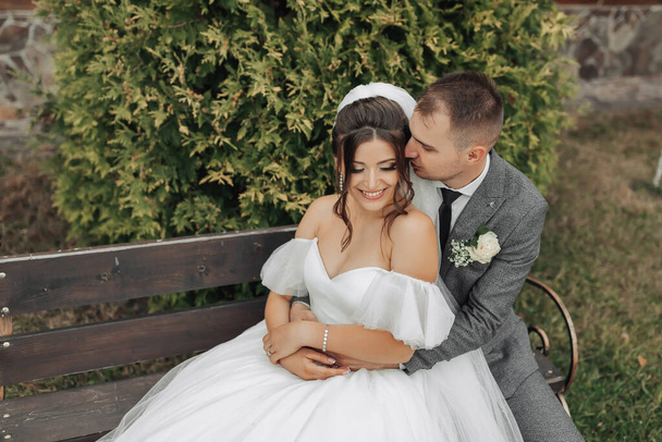 Portrait of the bride and groom in nature. A stylish groom and a brunette bride in a white long dress sit on a bench against a tree background, hug and sincerely smile. curly hair - Fotografie, Obrázek