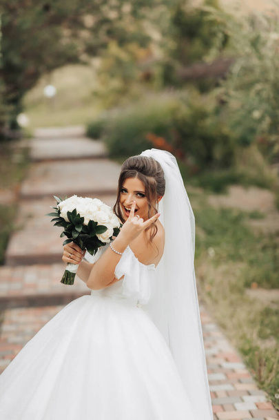 Portrait of the bride in nature. A brunette bride in a white long dress, holding a bouquet of white roses, poses, looking into the lens, smiling sincerely. on the background of the tree. Curly hair. - Photo, Image