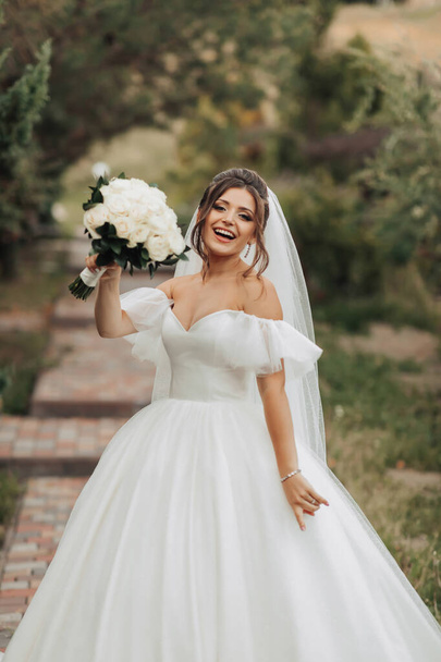 Portrait of the bride in nature. A brunette bride in a white long dress, holding a bouquet of white roses, poses, looking into the lens, smiling sincerely. on the background of the tree. Curly hair. - Foto, immagini