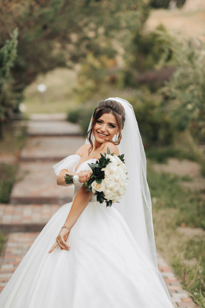 Portrait of the bride in nature. A brunette bride in a white long dress, holding a bouquet of white roses, poses, looking into the lens, smiling sincerely. on the background of the tree. Curly hair. - Foto, Imagen