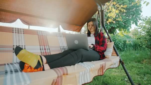Mladá dospělá žena pracující venku na zahradě s laptopem. Na volné noze pomocí notebooku, zatímco sedí na pohodlném houpačce na svém dvorku. Vysoce kvalitní 4K záběry - Záběry, video