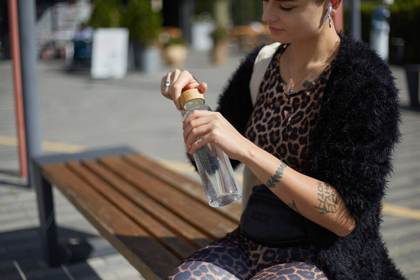 Thirsty young woman drinking water from a glass bottle outdoors. Fashionable female person opening a reusable bottle of water to drink. Healthy and sustainable lifestyle concept - 写真・画像