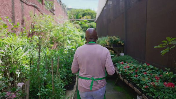Back of one black older person walking through garden in backyard of Flower Shop. Small business retail store of female African American employee - Photo, Image