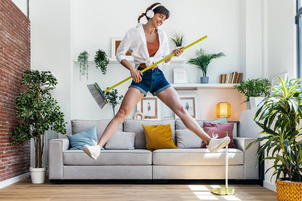 Shot of funny motivated woman dancing and listening to music with headphones while sweeping the house - Фото, изображение