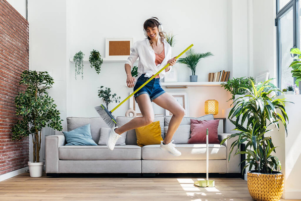 Shot of funny motivated woman dancing and listening to music with headphones while sweeping the house - 写真・画像