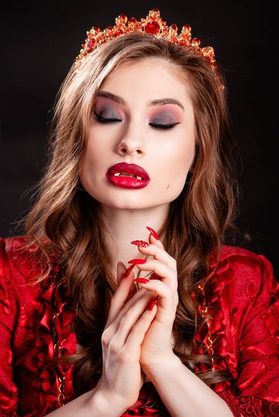 Portrait of a young, attractive vampire woman in a red dress in the rococo era with a crown on her head, posing isolated on a dark background. - Foto, Imagen