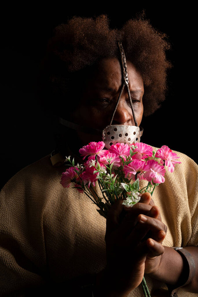 Πορτραίτο μιας μαύρης γυναίκας με αλυσίδες στα χέρια και μια σιδερένια μάσκα στο πρόσωπό της που κρατάει ένα μπουκέτο λουλούδια. Σκλάβος Αναστάσια. Δουλεία στη Βραζιλία. - Φωτογραφία, εικόνα