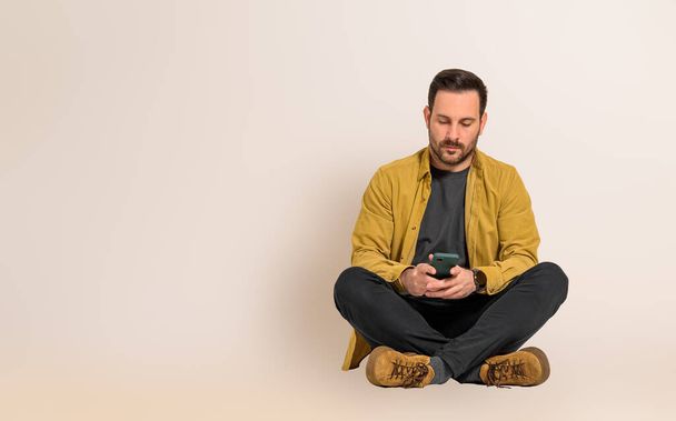 Σοβαρός νεαρός όμορφος επιχειρηματίας που διαβάζει μηνύματα στο κινητό ενώ κάθεται με σταυρωμένα τα πόδια. Άνδρας επαγγελματίας ντυμένος με περιστασιακή έλεγχο των μέσων κοινωνικής δικτύωσης στο κινητό με φόντο - Φωτογραφία, εικόνα