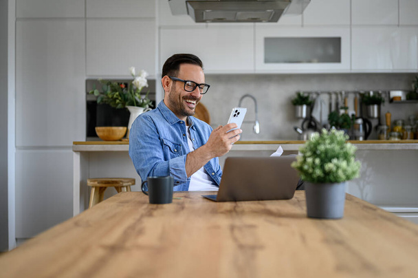 Alegre mensajería freelancer masculina a través del teléfono móvil con computadora portátil y café en el escritorio en la oficina en casa - Foto, Imagen