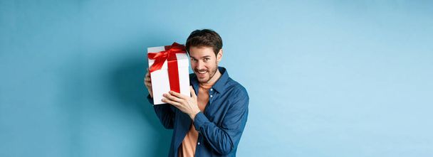 Ημέρα του Αγίου Βαλεντίνου. Ρομαντικό αγόρι δείχνει κουτί δώρου για τον εραστή, γιορτάζει την επέτειο, στέκεται πάνω από το μπλε φόντο. - Φωτογραφία, εικόνα