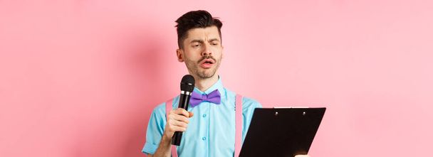 Концепция праздников. Серьёзно выглядящий мужчина читает сценарий из буфера обмена, держит микрофон, развлекает людей на праздничном мероприятии, стоя на розовом фоне. - Фото, изображение
