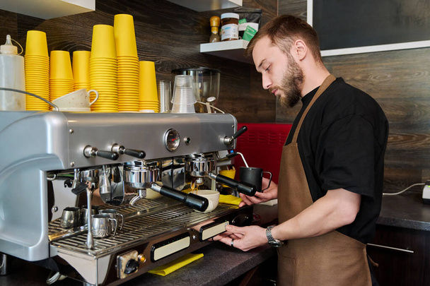 Male barista preparing coffee in coffee shop, bearded young man worker in an apron standing near coffee machine. Small business, service, coffee shop, staff, work concept - Foto, afbeelding