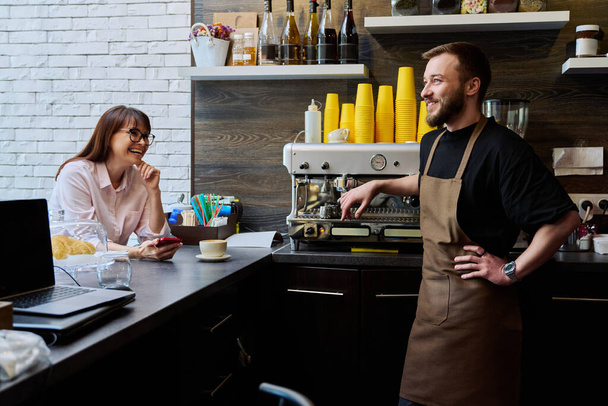 Young male barista talking to woman customer in coffee shop, near counter with cup of freshly prepared cappuccino coffee. Small business, service, coffee shop, staff, work concept - Foto, imagen