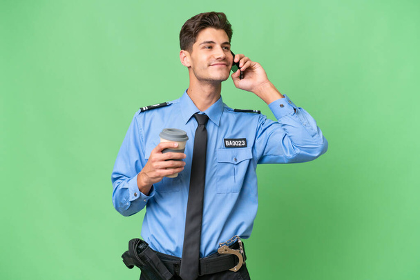 Młody policjant na odosobnionym tle, trzymający kawę do zabrania i komórkę. - Zdjęcie, obraz