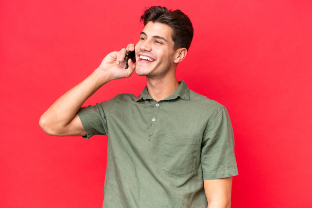 Jeune homme beau caucasien isolé sur fond rouge en gardant une conversation avec le téléphone mobile - Photo, image