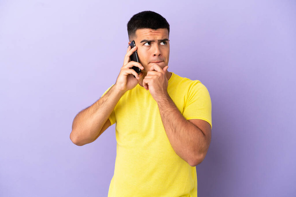 Mladý pohledný muž pomocí mobilního telefonu přes izolované fialové pozadí mají pochybnosti a myšlení - Fotografie, Obrázek