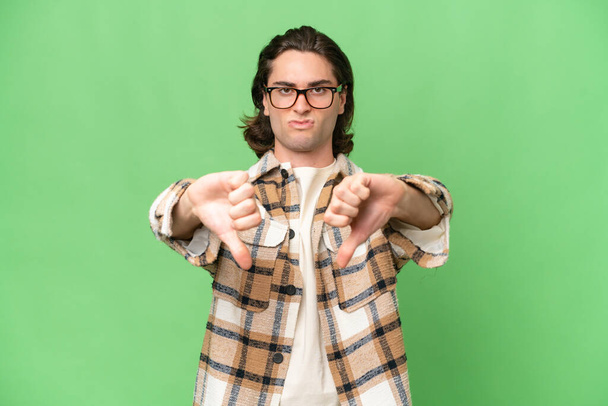 Young caucasian man isolated on green chroma background showing thumb down with two hands - Valokuva, kuva