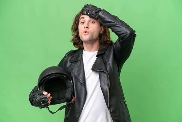 Joven hombre guapo con un casco de motocicleta aislado sobre fondo de croma verde haciendo gesto sorpresa mientras mira a un lado - Foto, imagen