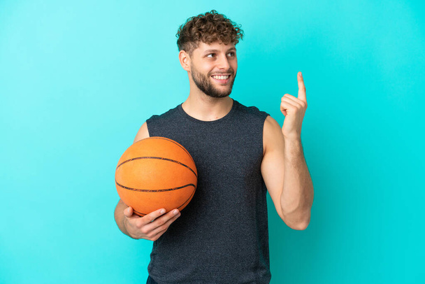 Beau jeune homme jouant au basket isolé sur fond bleu pointant vers le haut d'une excellente idée - Photo, image