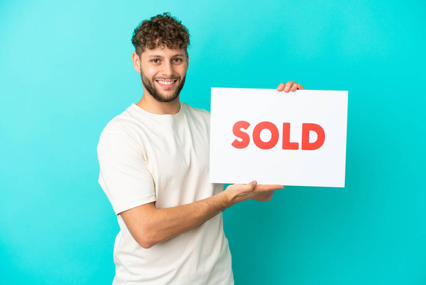 Young handsome caucasian man isolated on blue background holding a placard with text SOLD with happy expression - Photo, Image