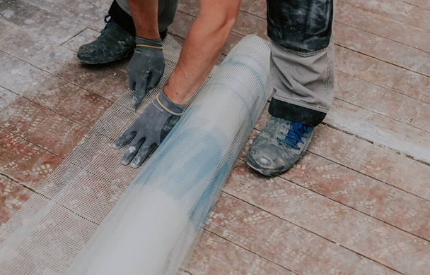 Junger kaukasischer, nicht wiedererkennbarer Mann in grauen Handschuhen schneidet mit einem Messer ein Baumaschinennetz auf einem schmutzigen Holzboden in einem Reparaturraum, Nahaufnahme von oben mit selektivem Fokus. Das Konzept der Renovierung von Häusern, Bauarbeiten. - Foto, Bild