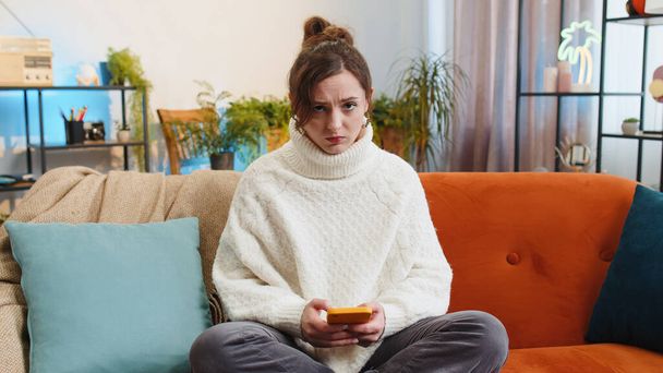 Sad displeased woman use smartphone typing browsing, loses becoming surprised sudden lottery results, bad news, fortune loss, fail, deadline, virus. Portrait of young girl at home living room on couch - Foto, Bild
