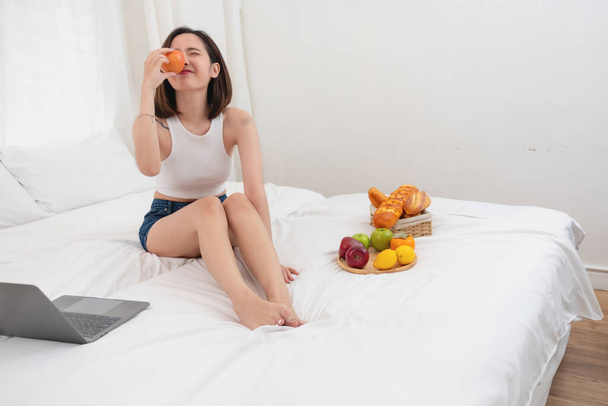 Retrato de mujer asiática blanca con un tatuaje sentado en la cama relajante y jugando portátil en fin de semana Había pan y frutas y manzanas rojas en una cama blanca para comer. concepto de vacaciones - Foto, imagen