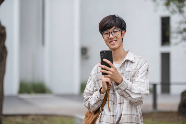 asiático macho universidad estudiante con inteligente teléfono en sunny campus. - Foto, imagen