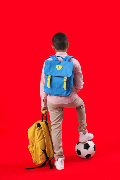 Little African-American schoolboy with soccer ball and backpacks on red background, back view - Photo, Image