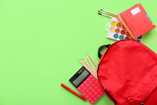 Roter Schulrucksack mit Taschenrechner, Farbstiften und Aquarellen auf grünem Hintergrund - Foto, Bild