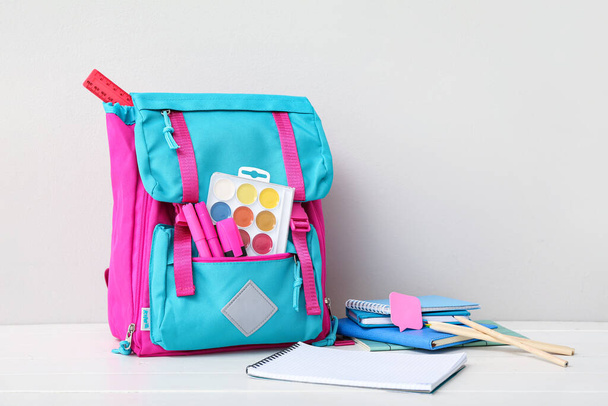 Рюкзак цвета школы с ноутбуками, акварелями и маркерами на белом деревянном столе у стены - Фото, изображение
