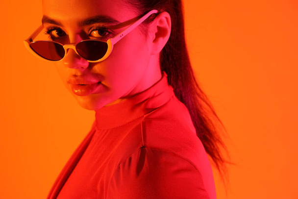 Fashionable young woman wearing sunglasses on orange background - Foto, Imagen