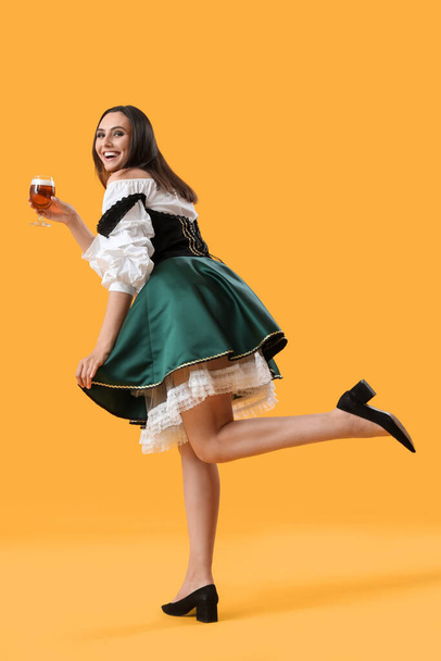 Beautiful Octoberfest waitress with beer dancing on yellow background - Photo, Image
