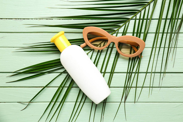 Okulary przeciwsłoneczne z butelką kremu do opalania i liściem palmy na zielonym drewnianym tle - Zdjęcie, obraz
