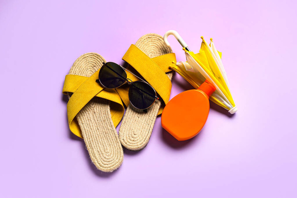 Occhiali da sole con infradito, mini ombrello e bottiglia di crema solare su sfondo lilla - Foto, immagini