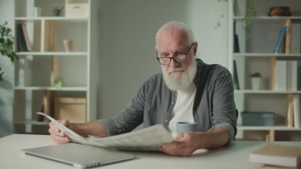 Ein ernsthafter, kluger alter Mann sitzt an einem Tisch und liest eine Zeitung. Ein älterer Mann mit einer Zeitung, der allein zu Hause Kaffee trinkt, sieht die Nachrichten und aktuellen Ereignisse in der Zeitung. - Filmmaterial, Video