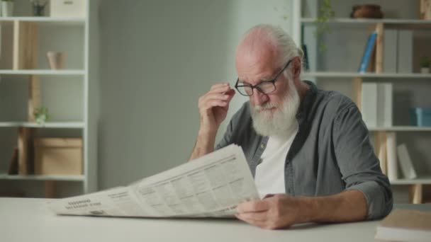 Een serieuze, slimme oude man zit aan een tafel en leest een krant.Een oudere man met een krant alleen thuis, ziet het nieuws en de huidige gebeurtenissen in de dagblad.Daily Newspaper Concept - Video