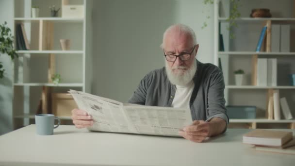 A Serious, Smart Old Manはテーブルに座り、新聞を読んでいます。自宅に一人で新聞を持っている高齢者は、新聞でニュースと現在のイベントを参照してください。日刊紙のコンセプト - 映像、動画