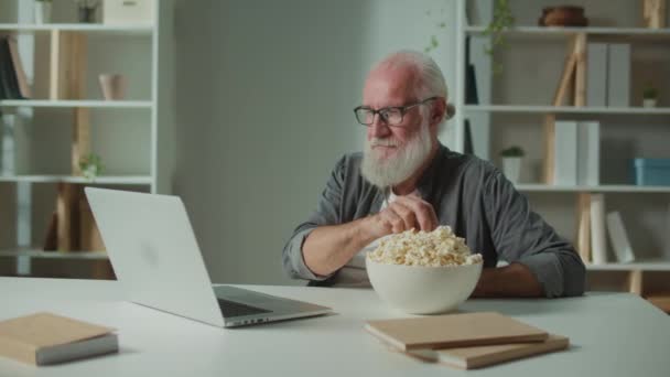 Улыбающийся старик смотрит фильм с ноутбука и ест попкорн. Веселый пожилой человек смеётся, смотря смешной фильм. Фильмы как форма релаксации и психологической помощи. - Кадры, видео