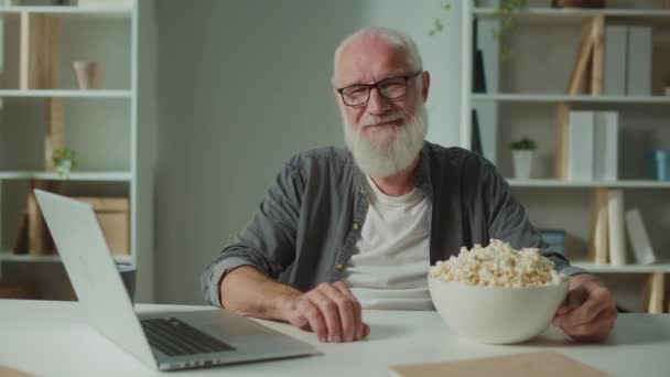Un ritratto di un vecchio sorridente nei bicchieri con computer portatile e popcorn. Allegro uomo anziano sta per guardare un film divertente. Film come forma di relax e sollievo psicologico. - Filmati, video
