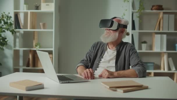 Un vecchio moderno con la barba grigia in VR occhiali esamina tutto intorno. Un uomo anziano con occhiali VR seduto a un tavolo con un computer portatile.Educazione alla realtà virtuale per anziani. - Filmati, video
