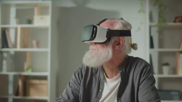 Un vecchio moderno con la barba grigia in VR occhiali esamina tutto intorno. Un uomo anziano con occhiali VR seduto a un tavolo a casa.VR per aiutare gli anziani. - Filmati, video
