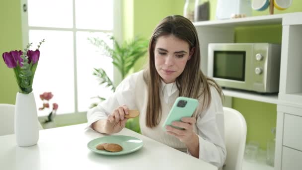 Νεαρή όμορφη ισπανόφωνη γυναίκα τρώει μπισκότα χρησιμοποιώντας smartphone στην τραπεζαρία - Πλάνα, βίντεο