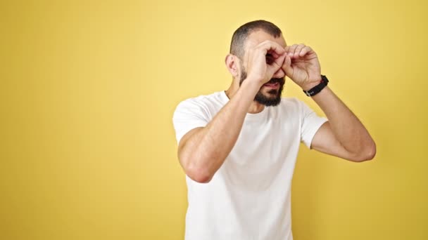 Joven hombre hispano haciendo binoculares gesto sonriendo sobre aislado fondo amarillo - Imágenes, Vídeo