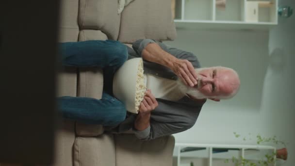 Vertikale Ansicht. Ein alter Mann isst Popcorn und schaut ein Sportprogramm. Ein ernsthafter älterer Mann sitzt auf dem Sofa und feiert einen Sportsieg. Filme als Form der Entspannung und psychologischen Entlastung. - Filmmaterial, Video